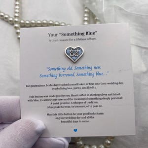 Può includere: Un bottone d'argento a forma di cuore con dettagli blu, con iniziali, poggia su una carta con testo a tema matrimonio. La carta recita "Something Blue" e include testo sull'amore e sui voti. Perle e decorazioni floreali circondano la carta.