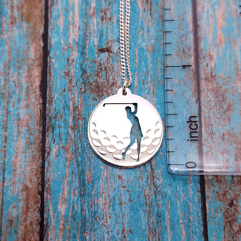 May include: Silver necklace with a round pendant featuring a golfer silhouette cut out against a textured golf ball design.