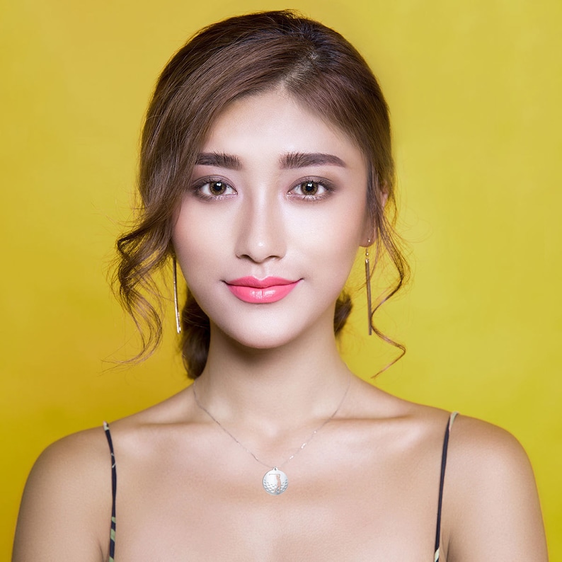 May include: A woman with long brown hair wearing a silver necklace with a round pendant and a pair of long, thin earrings. She is wearing a black spaghetti strap top and has a neutral expression on her face.