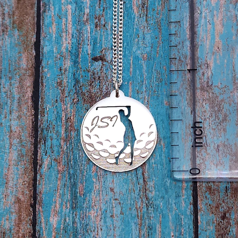 May include: Silver necklace with a round pendant featuring a golfer silhouette and a golf ball design. The pendant has the initials "JSJ" engraved on it.