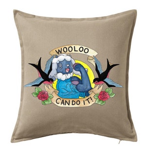 Op de afbeelding: Een beige kussen met een cartoon afbeelding van een blauw schaap met een zwart-wit gezicht. Het schaap buigt zijn biceps en heeft een banner boven zich met de tekst "Wooloo Can Do It!" De banner is versierd met rode rozen en zwarte zwaluwen.