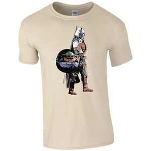 K&ouml;nnte beinhalten: Ein beiges T-Shirt mit einem grafischen Aufdruck eines Mandalorianer-Charakters aus Star Wars, der ein kleines gr&uuml;nes au&szlig;erirdisches Wesen h&auml;lt.