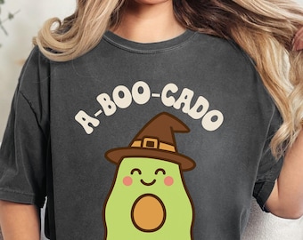 May include: Dark gray t-shirt with the text "A-BOO-CADO" above a cartoon avocado wearing a witch hat. The avocado is green with a brown pit and a smiling face. The shirt is a casual style.
