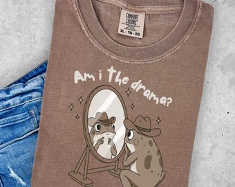 May include: Brown t-shirt with the text "Am I the drama?" and a cartoon frog wearing a cowboy hat looking in a mirror. The shirt is paired with blue denim jeans.