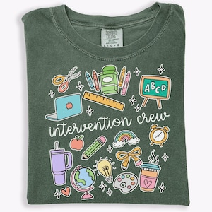 May include: A dark olive green t-shirt featuring a graphic print with the text "intervention crew" surrounded by colorful illustrations of school supplies including scissors, crayons, a ruler, a chalkboard, a laptop, an apple, a pencil, a globe, and a coffee cup.