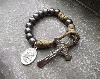 Op de afbeelding: Een zwart en groene paracord armband met een zilveren metalen kruis en een zilveren medaille met een religieus beeld. De armband ligt op een grijs, getextureerd oppervlak.