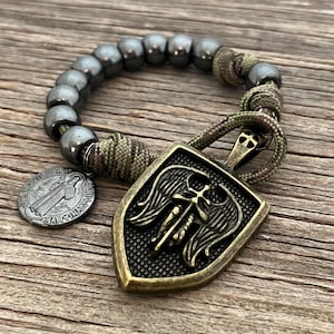 May include: A bracelet featuring dark gray beads and a shield-shaped pendant with an angel wing design. The pendant is attached to a camouflage cord. A small, round medallion with an inscription is also attached.