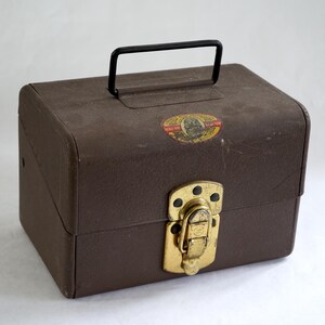 May include: A brown rectangular storage box with a black metal handle on top. The box features a gold-colored metal latch on the front and a small oval sticker with a dog illustration and text that reads "American Dog Food Co."