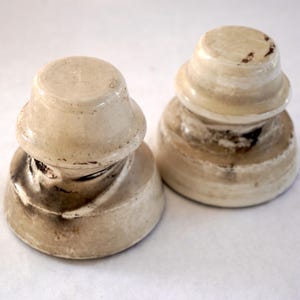 May include: Two ceramic insulators with a white glaze and brown markings. The insulators are stacked on top of each other.