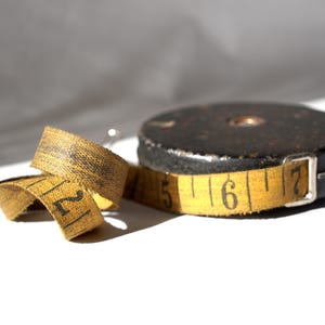 May include: A vintage yellow measuring tape with black numbers, wound around a black metal spool. The tape is marked with numbers 2, 5, 6, and 7.