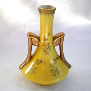 Könnte beinhalten: Eine gelbe Keramikvase mit goldenen Akzenten und Griffen. Die Vase hat ein goldenes Blumendesign und einen abgerundeten Boden. Die Oberseite und die Griffe sind goldfarben, und der Innenrand ist hellgoldfarben.