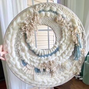 May include: A round woven wall mirror with a central circular mirror. The woven design features cream, white, and light blue yarns in various textures, including thick braids and fluffy sections. Beige fringe accents the design.