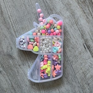 May include: A clear plastic unicorn-shaped bead organizer filled with colorful beads. The beads are in various shapes and sizes, including round, heart, and star shapes. The colors include pink, yellow, blue, and white.