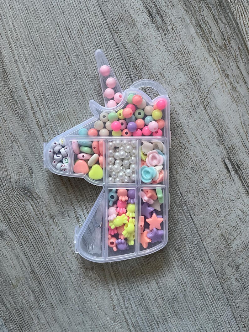May include: A clear plastic unicorn-shaped bead organizer filled with colorful beads. The beads are in various shapes and sizes, including round, heart, and star shapes. The colors include pink, yellow, blue, and white.