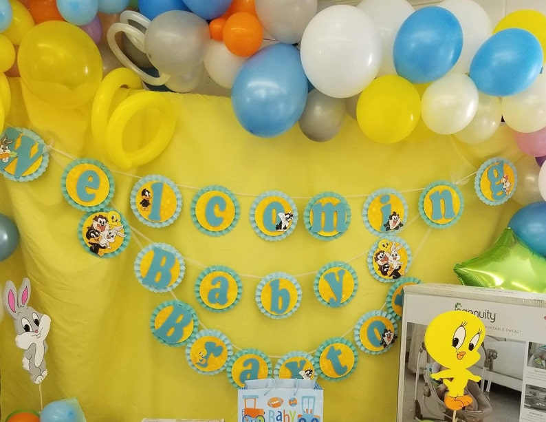 May include: A blue and yellow baby shower banner with the words "Welcome Baby" and cartoon characters. The banner is decorated with blue and yellow balloons.