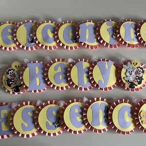May include: A pink and yellow baby shower banner with the words "Welcome Baby Essence" and cartoon characters from the Looney Tunes.