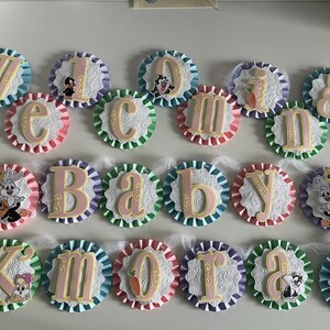 May include: A pink and white baby shower banner with the words "Welcome Baby Kmorah" in gold letters. Each letter is on a separate circle with a different cartoon character on each circle. The circles are connected with white tulle.
