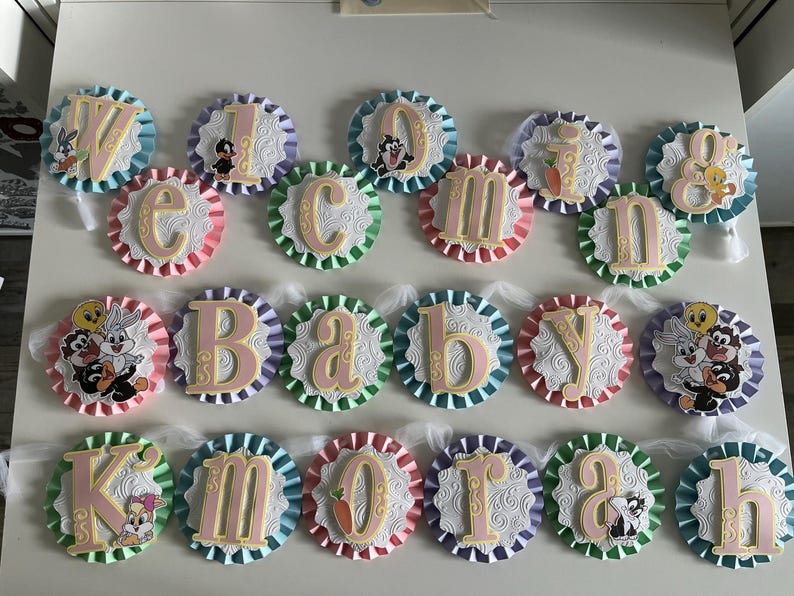May include: A pink and white baby shower banner with the words "Welcome Baby Kmorah" in gold letters. Each letter is on a separate circle with a different cartoon character on each circle. The circles are connected with white tulle.