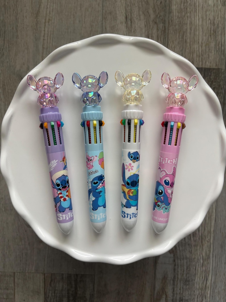 Op de afbeelding: Vier multi-gekleurde pennen met een cartoon karakter ontwerp. Elke pen heeft een verschillend gekleurde schacht, met een bijpassende topper. De pennen zijn gerangschikt op een wit, geschulpte bord. De pennen hebben het woord "Stitch" erop gedrukt.