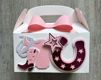 Puede incluir: Caja de regalo blanca con un lazo rosa y recortes con temática de vaquero. La caja presenta una herradura rosa con recortes de estrellas, una estrella, una bota de vaquero y un diseño de cuerda. El asa es blanca.