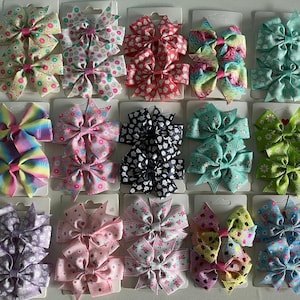 May include: A set of 10 pairs of colorful hair bows. The bows are made of ribbon and are arranged in rows on a white background. The bows are in various colors, including pink, blue, green, purple, and red.