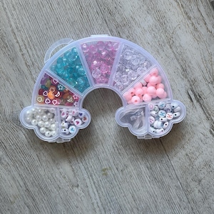 May include: A rainbow-shaped bead organizer filled with various colorful beads. The organizer is divided into compartments containing beads in shades of blue, pink, clear, and white. Some compartments have beads with letters and shapes.