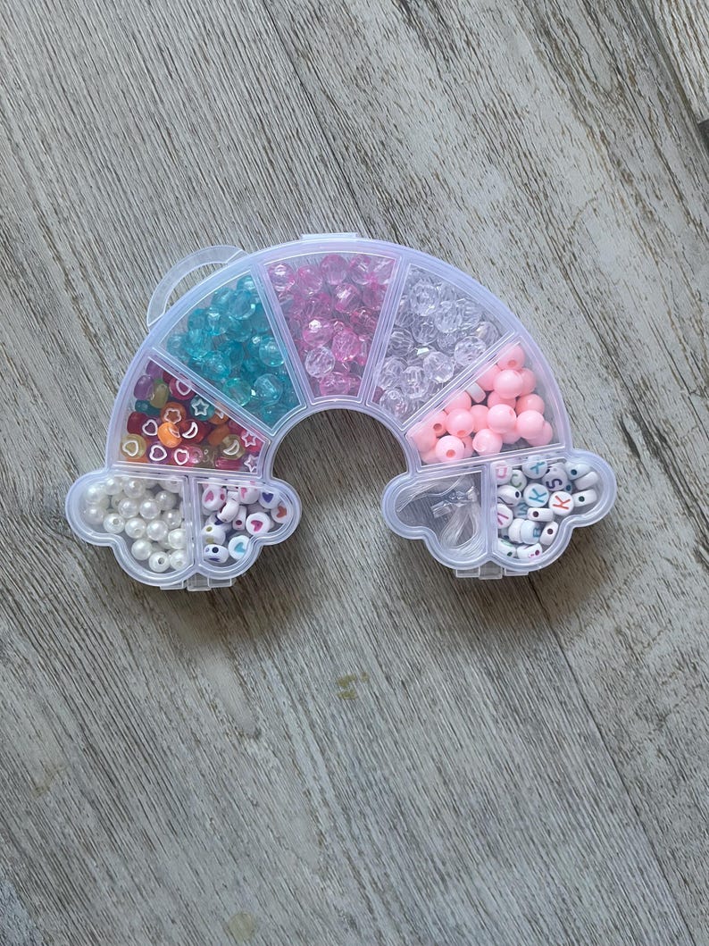 May include: A rainbow-shaped bead organizer filled with various colorful beads. The organizer is divided into compartments containing beads in shades of blue, pink, clear, and white. Some compartments have beads with letters and shapes.