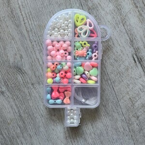 May include: A clear, popsicle-shaped container filled with various colorful beads and craft supplies. The container is divided into compartments holding pearls, pink beads, and assorted shapes like hearts and bows. A handle is on the side.