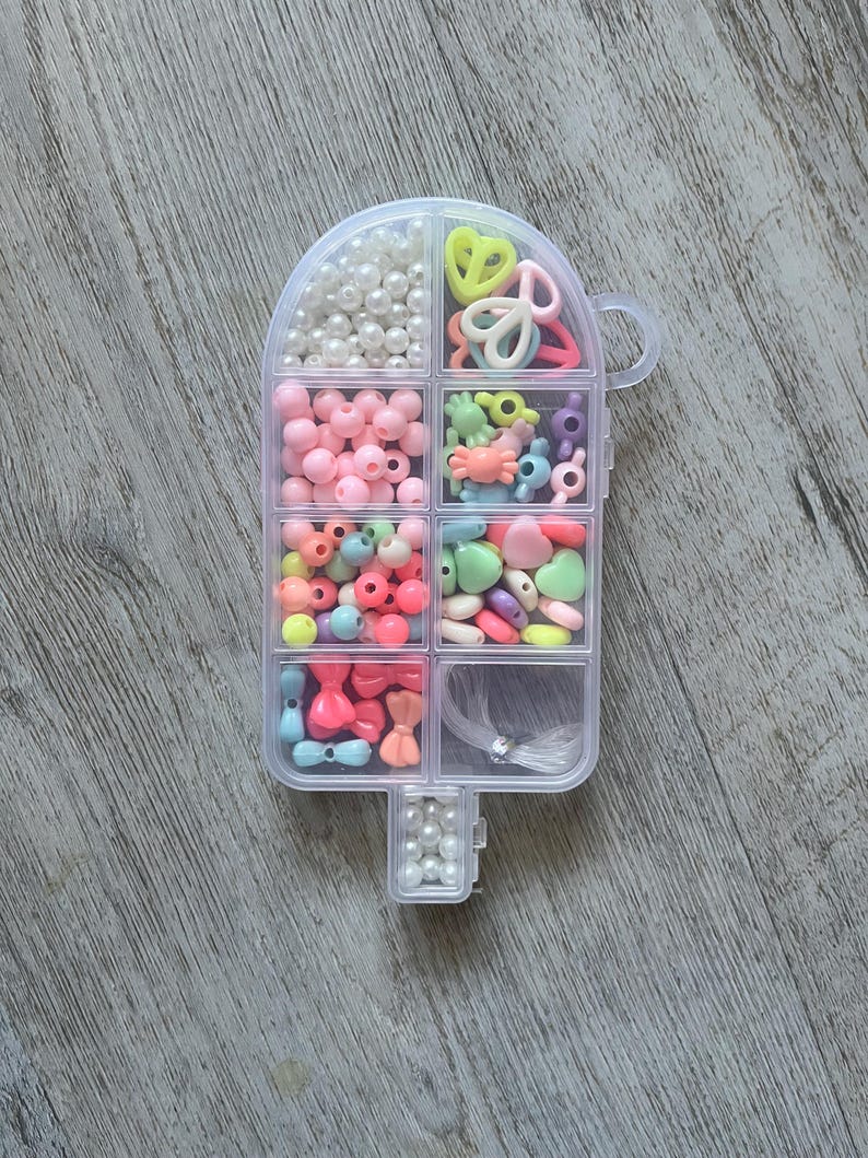 May include: A clear, popsicle-shaped container filled with various colorful beads and craft supplies. The container is divided into compartments holding pearls, pink beads, and assorted shapes like hearts and bows. A handle is on the side.