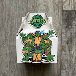 Peut inclure: Une boîte blanche à pignon avec un graphisme coloré des Tortues Ninja. La boîte porte un logo en forme de carapace de tortue verte avec le mot "Turtles" en blanc. La boîte est décorée avec les quatre tortues dans leurs couleurs emblématiques.