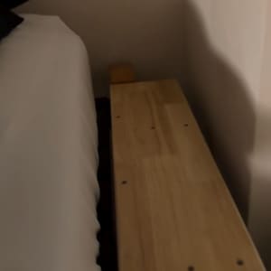 May include: A light-colored wooden bed frame with visible screw heads. The frame is positioned next to a white bed sheet and a wall. The wood has a natural grain and a smooth finish.