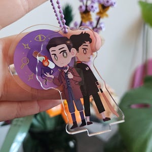 May include: A clear acrylic keychain featuring two cartoon characters. One character wears a brown coat and holds a book with a flame, while the other wears a black coat. The keychain has a purple chain and a purple circular design with stars and symbols.