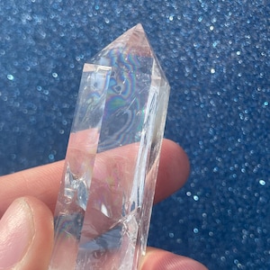 May include: A clear quartz crystal point with a faceted tip. The crystal is held against a sparkly blue background, and the light refracts through the quartz, creating a rainbow effect. The crystal is approximately 3 inches long.
