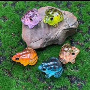 May include: A collection of five colorful frog figurines in shades of purple, green, orange, blue, and tan. The frogs are positioned on a rock and green grass, showcasing their vibrant colors and detailed designs.