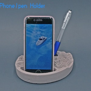 May include: A gray concrete phone and pen holder with a textured surface. A smartphone in a pink case is in the holder, and a silver pen with the text "Penn State University" is in the pen holder.