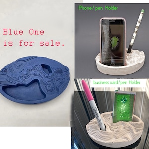 May include: Two concrete phone and pen holders. One is white and the other is blue. The white holder has a phone, pen, and business card in it. The blue holder is empty.