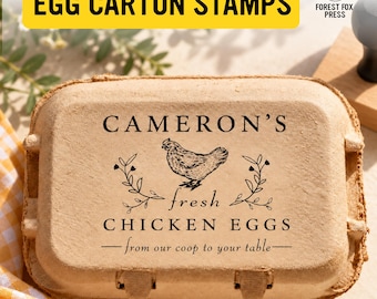 May include: A rectangular tan cardboard egg carton featuring a black ink stamp design. The stamp includes the text CAMERON'S, fresh, CHICKEN EGGS, and from our coop to your table, centered around a detailed illustration of a hen with floral accents.