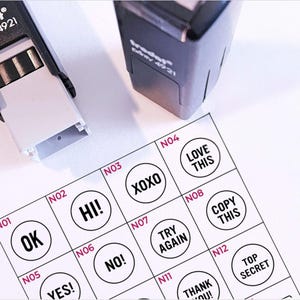 May include: A white sheet of paper with a grid of 12 circles containing different phrases. Each circle is numbered from 1 to 12. The phrases include "OK", "HI!", "XOXO", "LOVE THIS", "TRY AGAIN", "COPY THIS", "NO!", "YES!", "THANK YOU!", and "TOP SECRET".