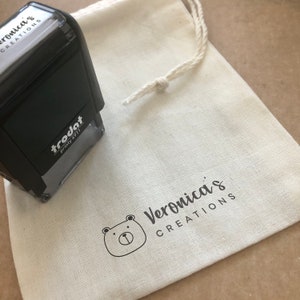 Peut inclure: Un sac en tissu blanc avec une fermeture &agrave; cordon. Le sac porte un logo noir et blanc qui dit "Veronica's Creations" avec une petite illustration d'ours. Un tampon d'encre noir avec le texte "trodat Printy 4911" est sur le dessus du sac.