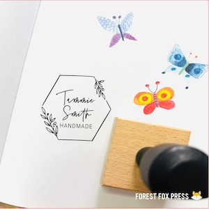 Peut inclure: Un tampon en bois avec un cadre hexagonal et le texte "Tannie Smith HANDMADE" à l'intérieur. Le tampon est utilisé pour créer une empreinte sur une surface blanche. Il y a trois illustrations de papillons aquarelle en arrière-plan.