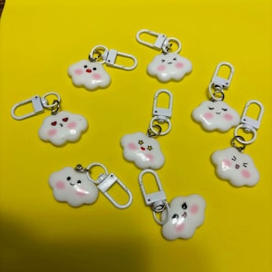 May include: A collection of white cloud-shaped charms, each featuring a unique facial expression with pink cheeks and a white metal lobster clasp attached to a small silver ring, scattered across the frame to display various designs.