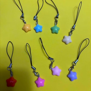 May include: Eight star-shaped charms in various pastel and bright colours, including orange, blue, green, white, red, pink, and purple. Each charm is attached to a thin black cord loop with a small silver-toned metal clasp and connector ring.