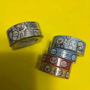 Op de afbeelding: Verschillende rollen decoratieve washi tape met cartoon illustraties. Eén rol toont een bureau scène met een persoon, terwijl andere verschillende gezichten en uitdrukkingen tonen. De tapes zijn in blauwe, rode en gele tinten.
