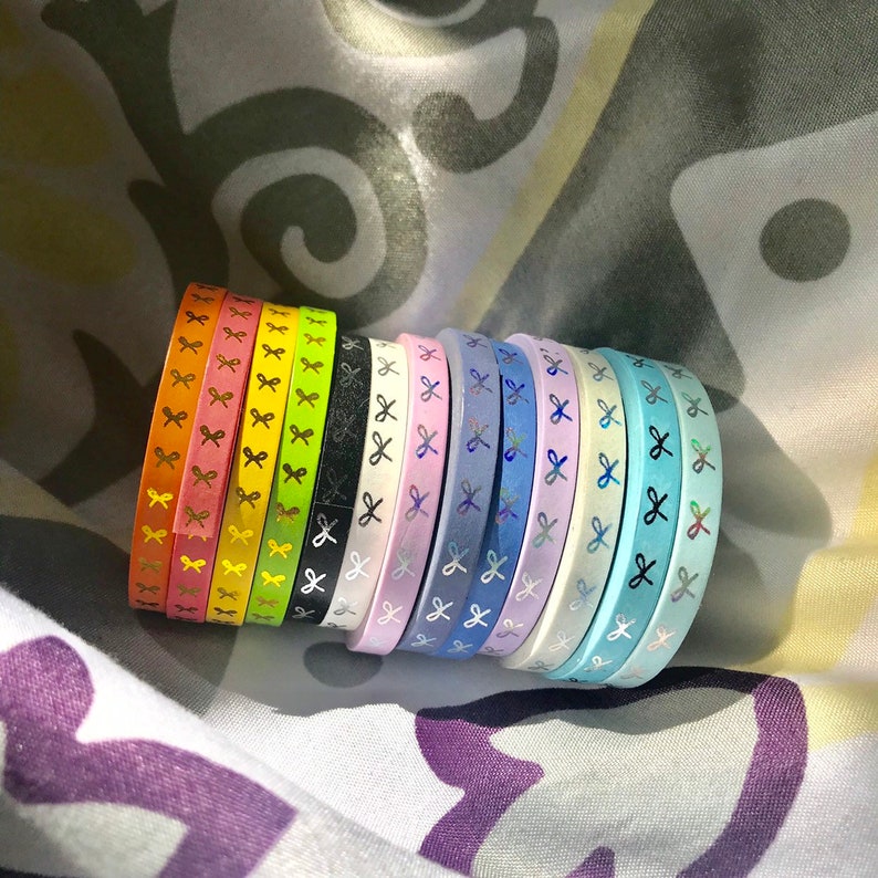 Op de afbeelding: Een verzameling kleurrijke washi tape rollen. Elke rol heeft een andere kleur, waaronder oranje, roze, geel, groen, zwart, wit, paars, blauw en turquoise. De tape is versierd met een herhalend strikpatroon.