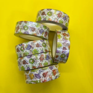 May include: A stack of patterned tape rolls featuring a repeating design of cartoon monsters in various colours. The tape has a white base with green, red, purple, and blue monster illustrations. The background is bright yellow.