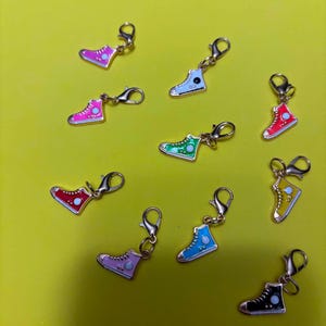 May include: A collection of nine metal trainer charms, each featuring a gold-tone lobster clasp. The charms are shaped like high-top trainers in various colours including pink, white, red, green, yellow, light purple, blue, and black with white details.