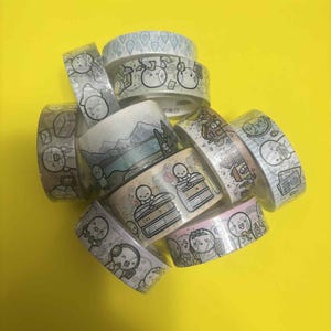 May include: A selection of decorative washi tape rolls with illustrated designs. The tape features images of people, books, landscapes, and other cute graphics. The rolls are stacked on a bright yellow background.