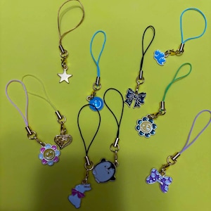 May include: A collection of nine phone charms featuring various metal and enamel pendants, including a gold star, a blue smiley face, a dark blue bow, a light blue cloud, a flower, a heart, a penguin, a rabbit, and a purple polka dot bow.