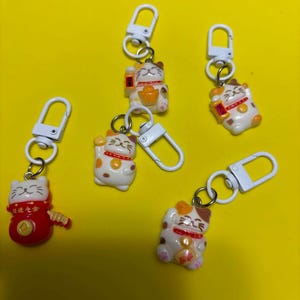 May include: Four small lucky cat keychains with white plastic lobster clasps. Three cats are white with brown and orange patches, and one is red. Each cat features a painted face, a collar, and holds a gold-coloured object. The red cat has text.