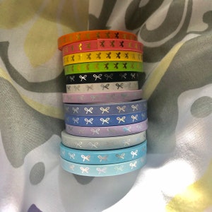 Op de afbeelding: Een stapel kleurrijke washi tape rollen. De rollen zijn in verschillende kleuren, waaronder oranje, roze, geel, groen, zwart, wit, lavendel, blauw en lichtblauw. Elke rol heeft een herhalend patroon van zilveren strikken.